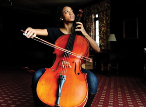 A black cellist, sitting down playing her cello, looking off to the side